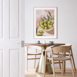 May include: A framed art print depicts a still life of green olives in a white bowl with olive branches. The artwork hangs above a round wooden table with a gold base, set with two cups and a creamer. Light wooden chairs surround the table.