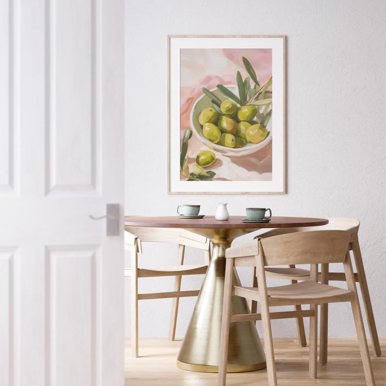 May include: A framed art print depicts a still life of green olives in a white bowl with olive branches. The artwork hangs above a round wooden table with a gold base, set with two cups and a creamer. Light wooden chairs surround the table.