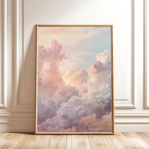 Op de afbeelding: Een verticale rechthoekige ingelijste wandkunstprint met een lucht vol zachte, volumineuze wolken in tinten pastelroze, lavendel en lichtblauw, verlicht door een warm, goudkleurig licht dat van achter de wolkenformaties vandaan komt.
