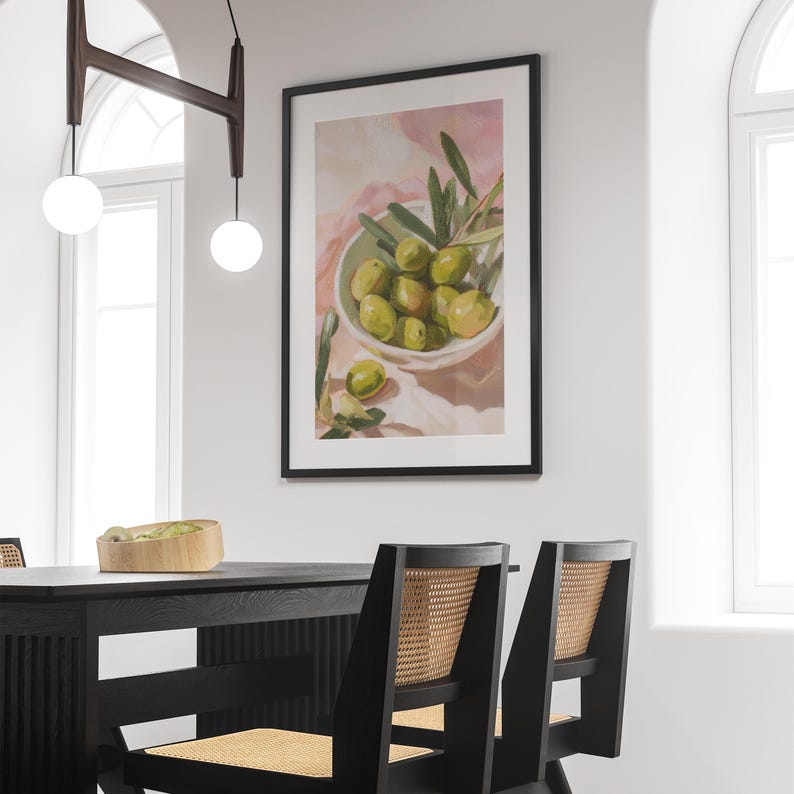 May include: A framed art print featuring a still life of green olives in a white bowl with olive branches. The artwork has a soft, painterly style with a pink and white background. The print is displayed on a white wall above a black dining table and chairs.