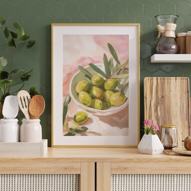 May include: A framed art print featuring a still life of green olives in a white bowl with olive branches. The painting has a gold frame and is set against a pink and white background. The artwork is displayed on a wooden cabinet.