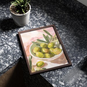May include: A framed art print featuring a still life of green olives in a white bowl, with olive branches. The artwork has a soft, painterly style with a brown frame. A small potted plant is in the background.