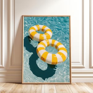 May include: A framed wall art print depicting two inflatable pool rings floating on clear blue water. The rings feature alternating yellow and white vertical stripes. The bright sunlight creates distinct, dark shadows of the rings on the pool floor.