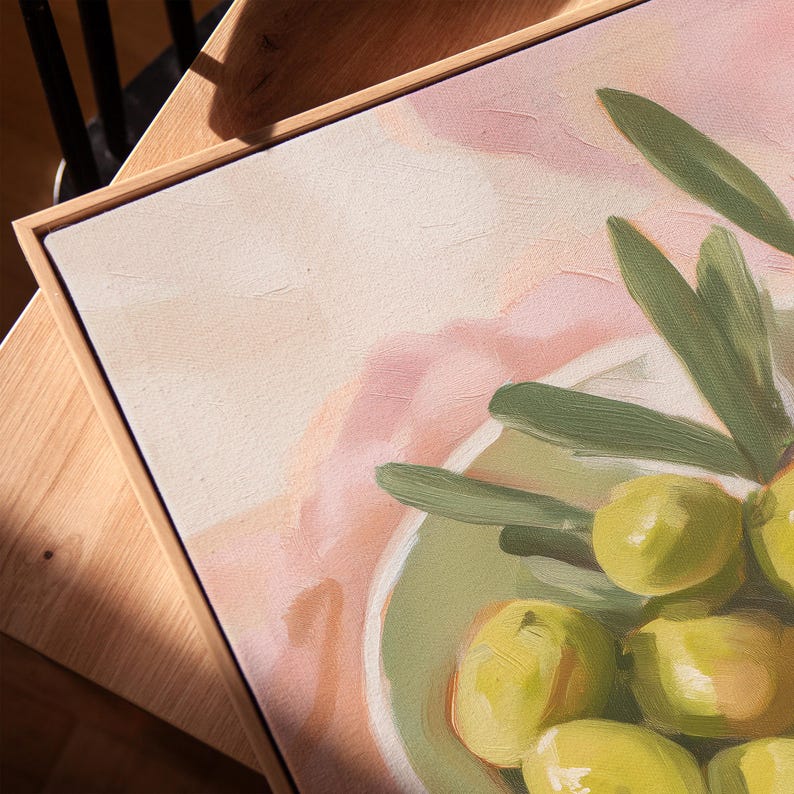 May include: Oil painting of green olives in a light green bowl, with olive branches. The artwork has a light pink and beige background, and is framed in a light wood frame. The painting is a close-up view.