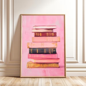 Puede incluir: Una impresión artística enmarcada que presenta una pila de libros en varios tonos de rosa, rojo y dorado. Los libros están dispuestos en forma de pirámide sobre un fondo rosa. La obra de arte está en un marco dorado.