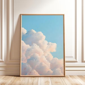 May include: A framed art print featuring a pastel painting of fluffy white and pink clouds against a light blue sky. The artwork is displayed in a light wood frame and leans against a white wall and wooden floor.
