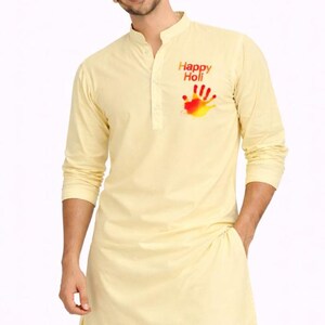 Può includere: Un kurta giallo chiaro con maniche lunghe e colletto alla coreana. Il kurta presenta un disegno con impronta di mano rossa, gialla e arancione con le parole "Happy Holi" stampate sopra. La persona indossa pantaloni bianchi.