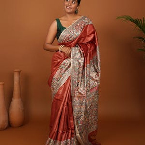 May include: A woman wearing a rust-colored sari with a detailed border and intricate artwork. The sari has a sheen and drapes elegantly. The woman is wearing a dark green top and gold jewelry.