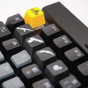 Puede incluir: Primer plano de un teclado mecánico negro con teclas personalizadas. Las teclas presentan varios iconos con temática de armas y una tecla amarilla brillante con el texto "CAS". El teclado tiene un diseño elegante y moderno.