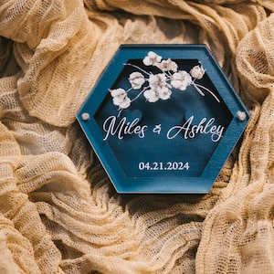 May include: A hexagonal blue ring box with a white floral design and the text "Miles & Ashley" and "04.21.2024".