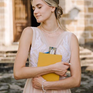 May include: A woman holding a yellow book with a bookmark. The woman is wearing a light pink dress with white embroidery and a pearl necklace and bracelet. The bookmark has the word "presa" on it.