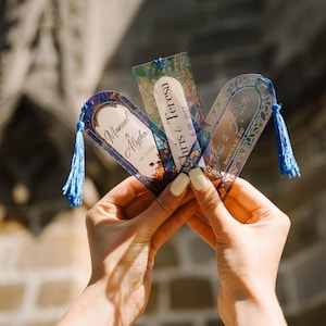 May include: Three personalized bookmarks with blue tassels. Each bookmark features a unique design with names and dates, including "Manuel & Alysha 09.13.2024", "Chris & Teresa 6.09.2027", and "Sofia & Adam 09.25.2027". The bookmarks have a stained glass effect.
