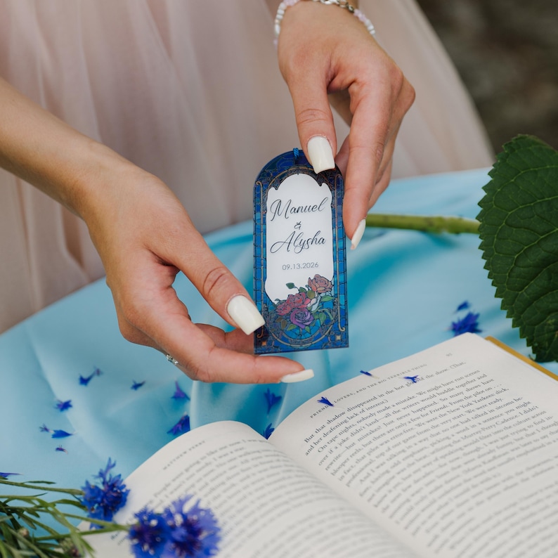 May include: A blue and gold wedding favor tag with the names "Manuel & Alysha" and the date "09.13.2026". The tag features a floral design with roses and a stained glass window effect. The tag is held in front of an open book.