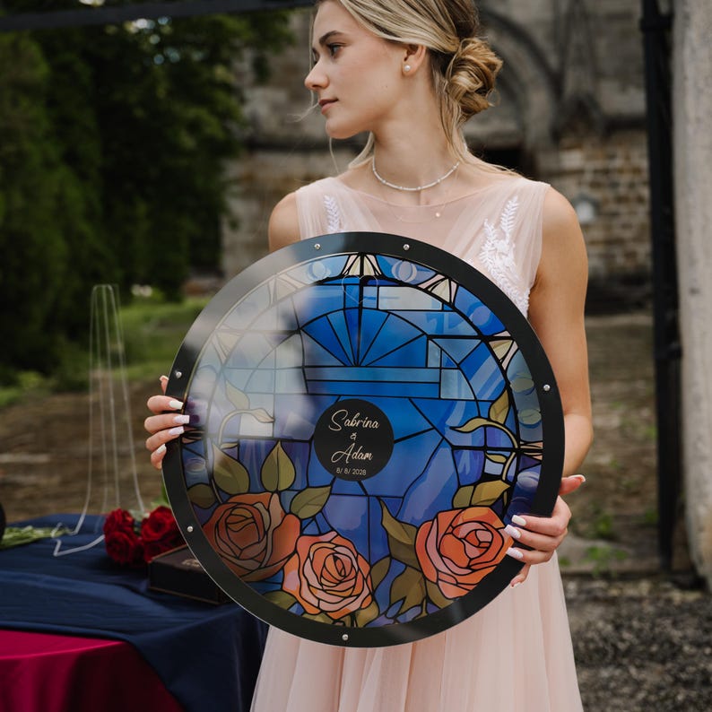 May include: A round, stained-glass-style wedding sign with a black frame. The design features a blue background with geometric patterns, orange roses, and the names "Sabrina & Adam" with a date. The sign is held by a person wearing a pink dress.