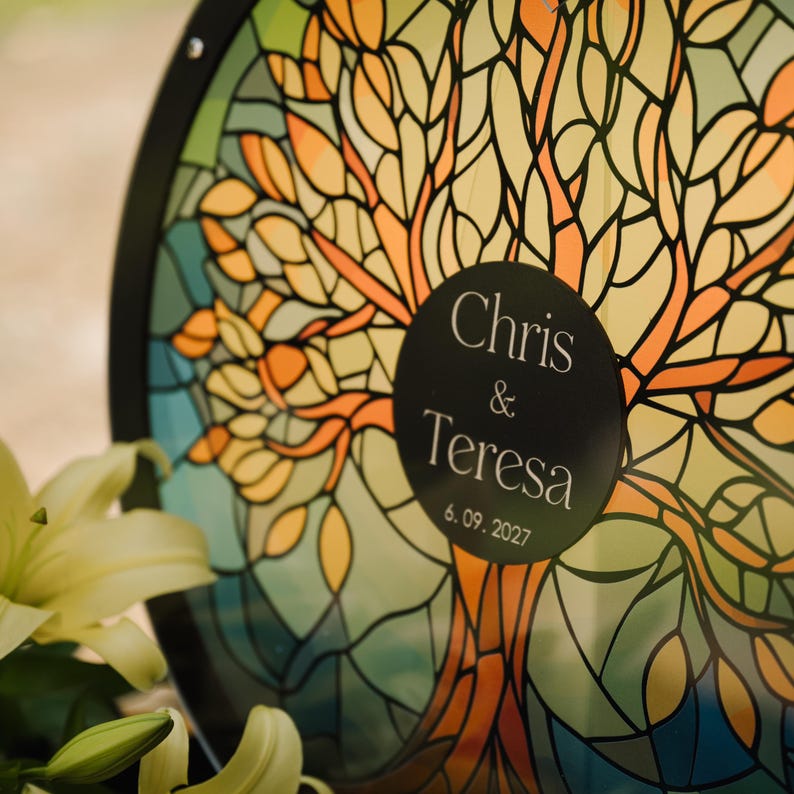 May include: A round, stained-glass style artwork featuring a tree design in shades of orange, yellow, and green. A black circular label displays the names "Chris & Teresa" and the date "6.09.2027". The artwork is framed in black.