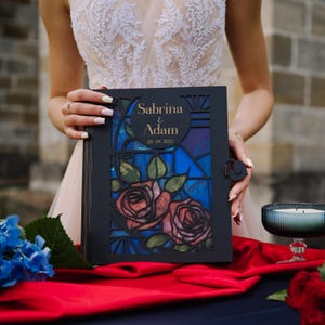 Peut inclure: Livre d'or de mariage noir avec un motif de vitrail représentant des roses et les noms "Sabrina & Adam" avec la date "09.09.2027". Le livre a un fermoir décoratif et est présenté sur un fond rouge et bleu.