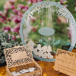 以下が含まれることがあります： 葉と枝の切り抜きデザインが施された木製の箱。箱は、小さな白いハート型の木片でいっぱいです。箱の上に、花柄が描かれた透明な丸い看板がぶら下がっています。看板には、黒い筆記体で「Allison & Craig」と書かれた小さな白い楕円形のラベルが付いています。看板は、手書きのメッセージが書かれた小さな白いハート型の木片でいっぱいです。画像の右側には、「ハートにサインをして箱に入れてください。特別な日の甘い思い出をあなたと一緒に大切にしましょう。」というテキストが書かれた看板があります。