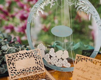 Peut inclure: Une boîte en bois avec un motif découpé de feuilles et de branches. La boîte est remplie de petits morceaux de bois en forme de cœur blancs. Un panneau rond transparent avec un motif floral peint est suspendu au-dessus de la boîte. Le panneau a une petite étiquette ovale blanche avec le texte "Allison & Craig" écrit en cursive noire. Le panneau est rempli de petits morceaux de bois en forme de cœur blancs avec des messages manuscrits dessus. Un panneau avec le texte "Signez un cœur et déposez-le dans la boîte. Gardons de doux souvenirs de notre journée spéciale avec vous." est sur le côté droit de l'image.