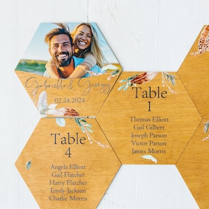 Op de afbeelding: Houten zeshoekige tafelnummerkaarten met bloemmotieven en de namen van de gasten. De bovenste kaart toont een foto van een stel en de tekst "Gabriello & Georgiy 02.24.2024".