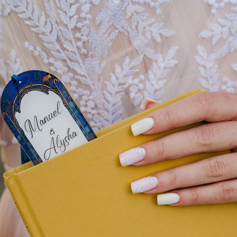 May include: A personalized bookmark with the names "Manuel & Alysha" in elegant script, inserted into a yellow book. The bookmark features a stained-glass design with blue and gold accents. The background shows a white lace fabric.