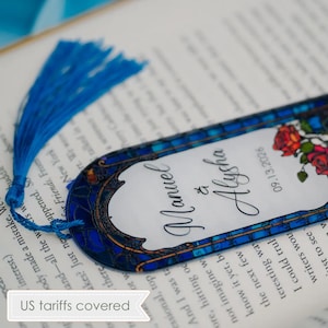 May include: A decorative bookmark with a blue tassel, featuring a stained-glass design. The bookmark has the names "Manuel & Alysha" and the date "09.13.2026" printed on it, along with a floral motif. The bookmark is resting on an open book.
