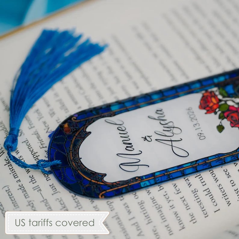 May include: A decorative bookmark with a blue tassel, featuring a stained-glass design. The bookmark has the names "Manuel & Alysha" and the date "09.13.2026" printed on it, along with a floral motif. The bookmark is resting on an open book.