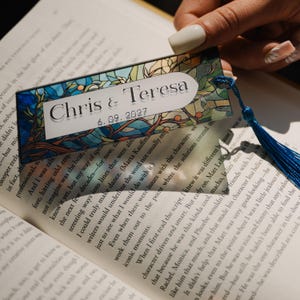 May include: A rectangular bookmark with a stained-glass design featuring blue, green, and orange hues. The bookmark has the names "Chris & Teresa" and the date "6.09.2027" printed on it. A blue tassel hangs from the bottom.