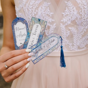 May include: Three personalized bookmarks with blue tassels. Each bookmark features a different name and date, with floral and stained glass designs. The names are Manuel & Alysha, Chris & Teresa, and Sabrina & Adam.