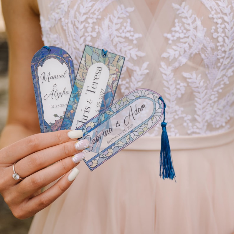 May include: Three personalized bookmarks with blue tassels. Each bookmark features a different name and date, with floral and stained glass designs. The names are Manuel & Alysha, Chris & Teresa, and Sabrina & Adam.