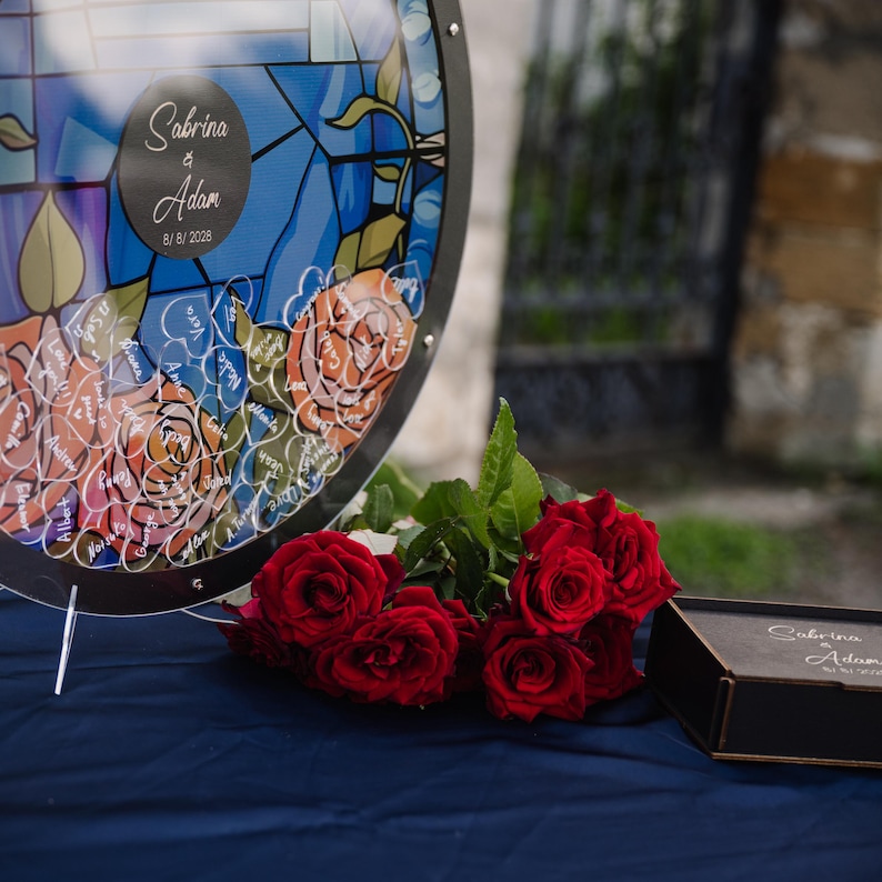 May include: A round, stained-glass-style wedding guest book with the names "Sabrina & Adam" and the date "8/8/2026". The book features rose designs and is accompanied by red roses and a small black box.