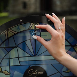 May include: A hand holding a heart-shaped acrylic piece with the word "Love" on it, in front of a stained-glass-style design. The design features blue and green hues, with the name "Sabrina" visible at the bottom.