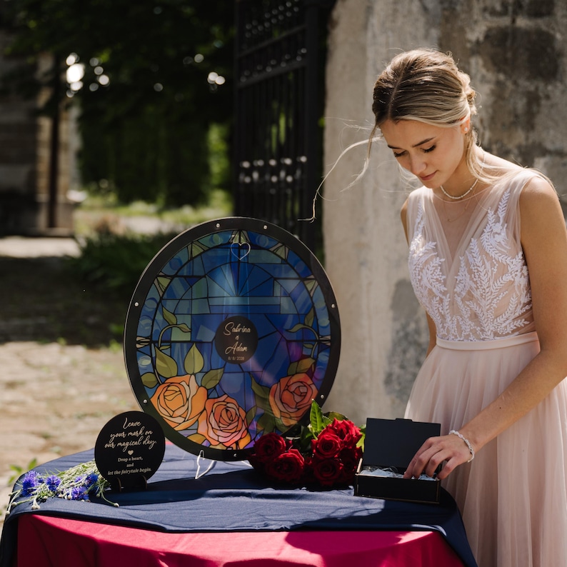 May include: A round, stained-glass-style wedding sign with a blue background and orange roses. The sign has the names "Sabrina & Alan" and a date. A small black sign reads "Leave your mark on our magical day". A woman in a wedding dress stands nearby.