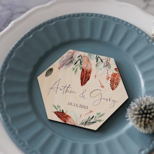 May include: A hexagonal wooden wedding place card with a watercolor design of feathers and leaves in shades of brown, orange, and green. The text "Anthea & George" and the date "10.15.2023" are printed in purple.