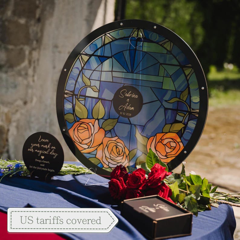 May include: A round, stained-glass-style wedding sign featuring a blue and green design with orange roses. The sign includes the names "Sabrina & Adam" and the date 8/2028. A smaller sign reads "Leave your mark on our magical day."