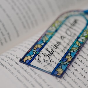 May include: A clear, stained-glass style bookmark with the name "Sabrina" in elegant script. The bookmark features a blue border and colorful floral and geometric designs. It rests on the open pages of a book.