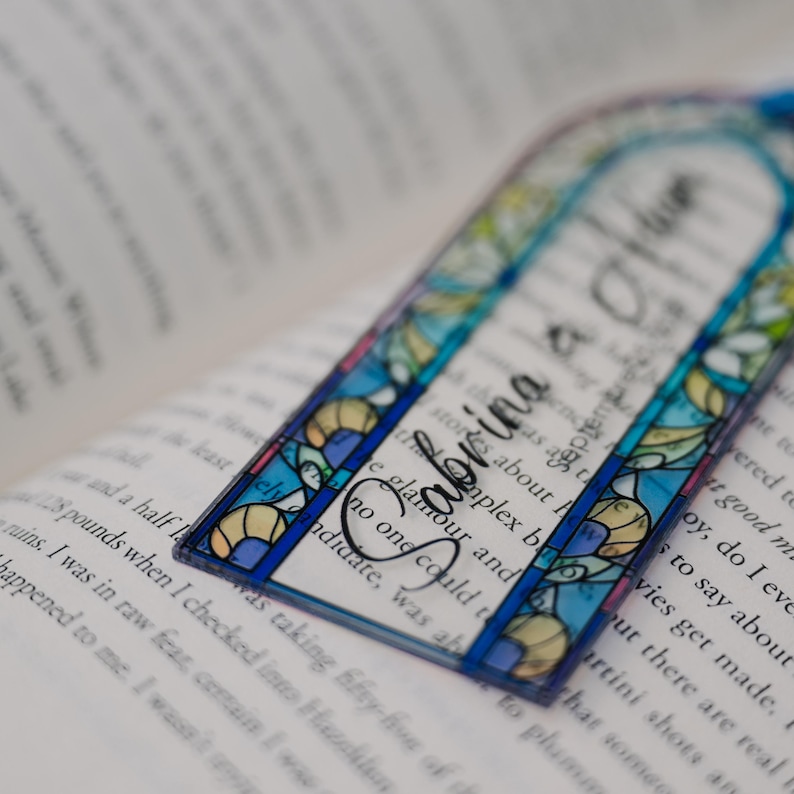 May include: A clear, stained-glass style bookmark with the name "Sabrina" in elegant script. The bookmark features a blue border and colorful floral and geometric designs. It rests on the open pages of a book.