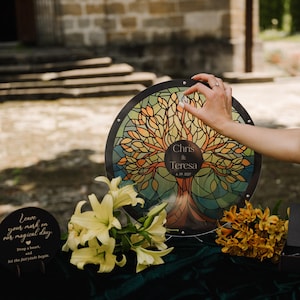 May include: A round, stained-glass style wedding guest book alternative featuring a tree design with the names "Chris & Teresa" and the date "6.09.2007". The design is set against a black border. The image also includes a sign that says "Leave your mark on our magical day."