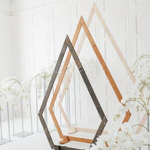 Könnte beinhalten: Drei geometrische Hochzeitsbögen aus Holz in verschiedenen Braun- und Beigetönen stehen vor einer weißen Wand. Weiße Blumengestecke sind auf beiden Seiten der Bögen platziert. Die Bögen sind aus abgewinkelten Holzbalken gefertigt.