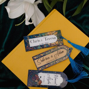 May include: Three personalized bookmarks with blue tassels rest on a yellow book. The bookmarks feature names and dates: "Chris & Teresa 6.09.2027", "Sabrina & Adam September 25, 2027", and "Manuel & Alysha 09.13.2026".