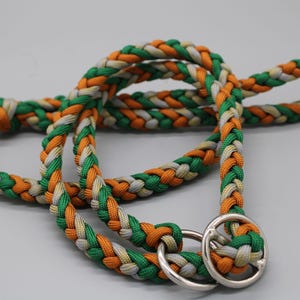 Könnte beinhalten: Geflochtene Hundeleine in Grün, Orange und Weiß. Die Leine besteht aus einem dicken, strapazierfähigen Material und hat am Ende einen silbernen Metallring. Die Leine ist geschlungen und liegt auf einer weißen Oberfläche.