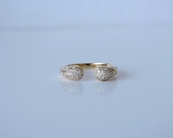 May include: A gold-colored open ring with two rounded ends, each encrusted with small, sparkling clear stones. The ring is set against a light, reflective surface, highlighting its polished finish and intricate design.