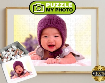 Może przedstawiać: Personalizowane puzzle ze zdjęciem uśmiechniętego niemowlęcia w fioletowej czapce z dzianiny i szarym ubranku. Puzzle są umieszczone w drewnianej ramie, obok znajduje się metalowe pudełko z elementami układanki oraz wydruk zdjęcia.