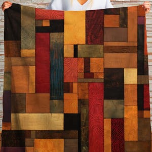 May include: A patchwork quilt featuring a geometric design of rectangular and square shapes in various colors. The quilt showcases a mix of textures and hues, including red, orange, brown, black, and gold. The quilt is held up by a person.