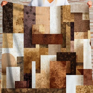 May include: A geometric patterned blanket featuring a collage of rectangular shapes in shades of brown, beige, and white. The design has a textured appearance, creating a modern and abstract aesthetic. The blanket is held up by a person.
