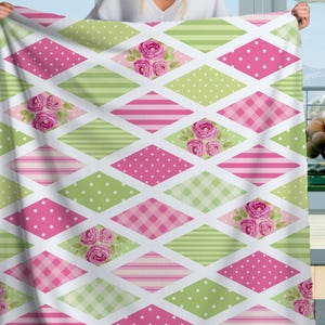 Pode incluir: Um cobertor decorativo com um padrão de losangos. O design inclui bolinhas cor-de-rosa, bolinhas verdes, listras cor-de-rosa, listras verdes e xadrez rosa. Acabamentos florais de rosas também estão incluídos. O cobertor é segurado.