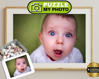 Könnte beinhalten: Ein individuelles Puzzle mit einer Nahaufnahme eines Babys mit großen blauen Augen und offenem Mund vor grünem Hintergrund. Das Puzzle ist in einem Holzrahmen gerahmt, daneben steht eine Metalldose mit losen Puzzleteilen.