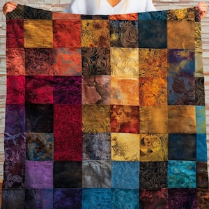 Puede incluir: Un edredón de retazos con cuadrados texturizados en tonos de rojo, morado, dorado, azul y marrón. El edredón se muestra contra una pared de ladrillos claros, mostrando sus colores vibrantes y patrones intrincados.