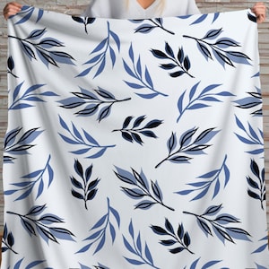 May include: White blanket with a repeating pattern of blue and black leaf branches. The leaves are stylized with a modern, minimalist design. The blanket is held up against a brick wall.