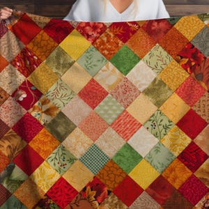 Könnte beinhalten: Ein farbenfrohes Patchwork-Quilt mit einem Rautenmuster. Der Quilt zeigt eine Vielzahl von Stoffen in Rot-, Orange-, Gelb-, Grün- und Cremetönen. Das Design umfasst Blumen- und Blattmuster und schafft eine warme und einladende Ästhetik.