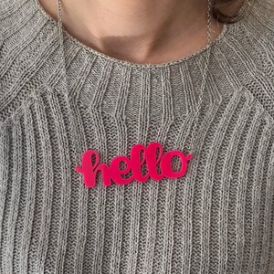 Könnte beinhalten: Eine silberne Kettenkette mit einem leuchtend pinkfarbenen Acryl-Anhänger, auf dem in kursiv "hello" steht.